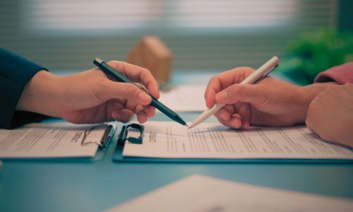 Close-up of two people reviewing and signing a document on a clipboard, each pointing with a pen at sections of the paperwork, suggesting a formal agreement or contract discussion.