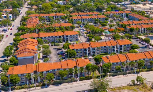 Aerial view of a dense residential neighborhood with rows of low-rise apartment buildings with orange tile roofs, tree-lined streets, and surrounding urban development.