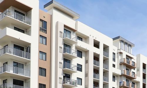 A modern mid-rise apartment building with multiple balconies, large windows, and a mix of white, beige, and brown exterior finishes under a clear blue sky.