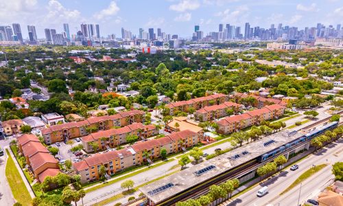 Aerial view of a Miami neighborhood with rows of low-rise apartment buildings surrounded by trees, a Metrorail line running alongside a main road in the foreground, and the downtown Miami skyline visible in the distance under a clear sky.