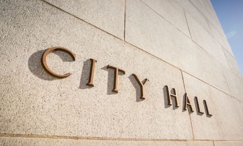The image shows the words "City Hall" engraved in large, metallic letters on a textured stone wall, likely the exterior of a city government building. The letters cast subtle shadows on the surface, suggesting daylight. The design is simple and formal, reflecting the significance of the building