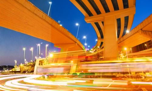Illuminated highway overpasses intersect at night, with sweeping concrete pillars and light trails from passing traffic.