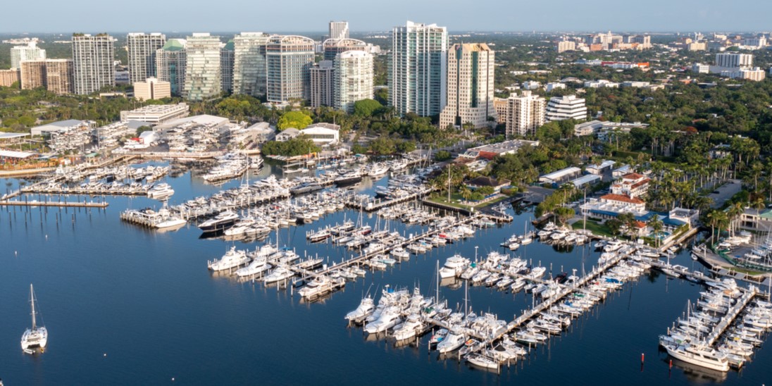 Aerial view of the Coconut Grove marina filled with boats beside high-rise buildings and lush greenery.