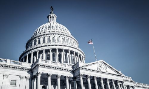 Exterior view of the United States Capitol building with the American flag flying in the foreground against a clear sky. 