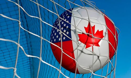 Soccer ball featuring U.S. and Canadian flag designs caught in a goal net against a clear blue sky.