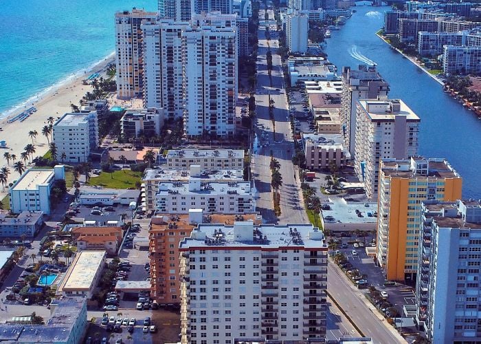 Aerial view of a coastal cityscape with high-rise buildings lining both the beach and an inland waterway, separated by a central road.