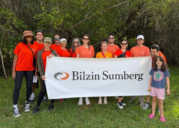 Group of Bilzin Sumberg volunteers in orange shirts pose holding a banner by the Mangrove Preserve during a community cleanup event in North Miami Beach.