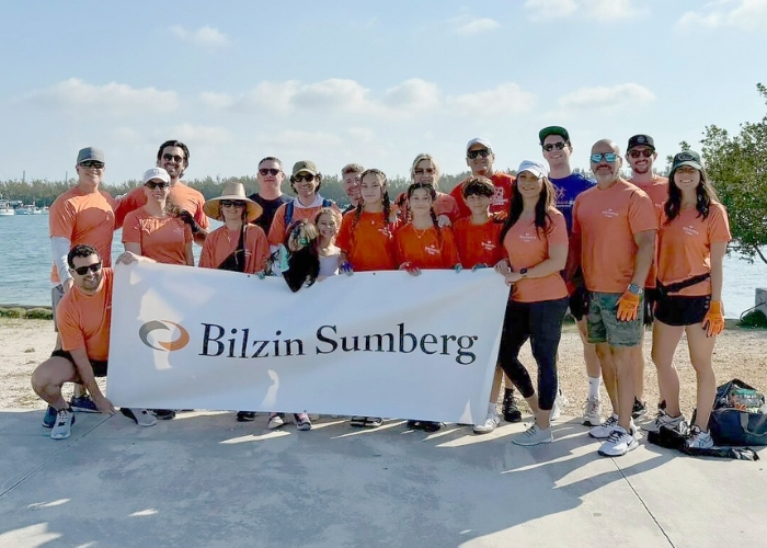 Bilzin Sumberg volunteers in orange shirts hold a banner by the waterfront during a community cleanup event on a sunny day.