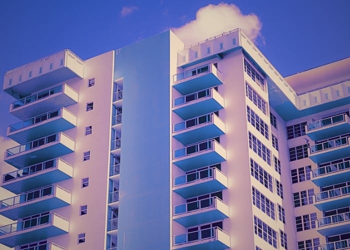 Pastel-toned high-rise apartment building with stacked blue balconies against a vivid sky.